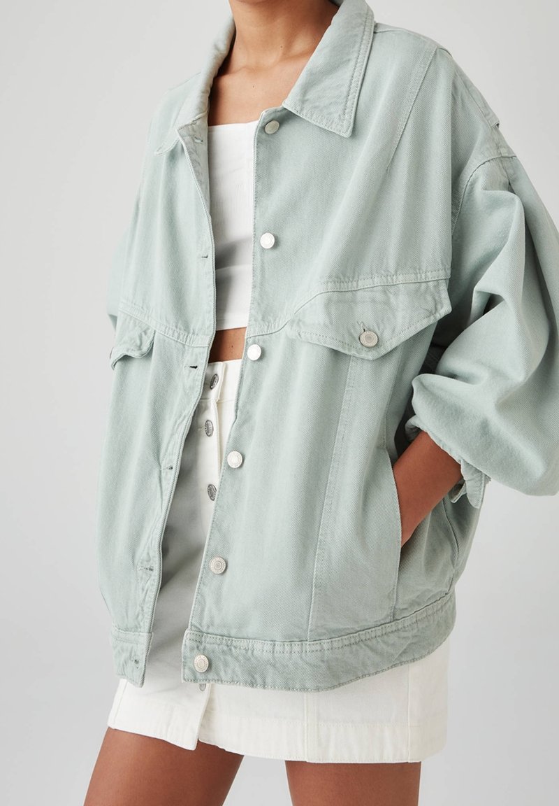 Veste en denim oversized vert clair avec un boutonnage à l'avant, deux poches poitrine et une finition légèrement textured, portée sur un haut blanc.