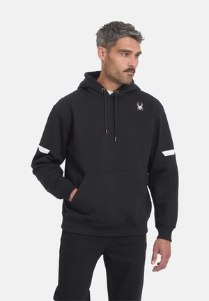 Mann mit kurzem grauem Haar, der einen schwarzen Hoodie mit weißem Spinnenlogo auf der Brust und weißen Streifen an den Ärmeln trägt, Hand in der Tasche, blickt nach rechts.