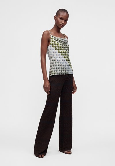 Mannequin féminin portant un haut sans manches vert et blanc à motifs et un pantalon noir à jambes larges avec des sandales ouvertes, debout sur fond blanc.