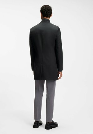 Homme debout de dos, portant un long manteau noir, un pantalon gris et des chaussures noires sur un fond blanc uni.
