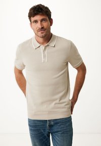 Beige polo triko vyrobené z měkkého pleteného materiálu, s klasickým límečkem, tříknoflíkovou légou a kontrastními bílými detaily na manžetách.
