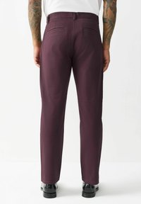 Bordowe spodnie o prostym kroju, z dwiema tylnymi kieszeniami i gładką fakturą. Noszone z czarnymi butami, widok z tyłu.