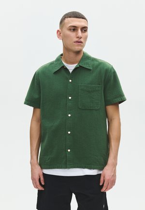 Mand iført en struktureret, kortærmet, grøn skjorte med knapper over en hvid T-shirt og sorte shorts, stående mod en ensfarvet hvid baggrund.