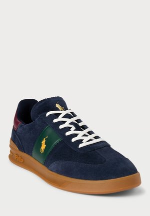 Granatowy zamszowy sneaker z białymi sznurowadłami, zielonym panelem bocznym z żółtym logo Polo, bordowym obcasem i brązową gumową podeszwą.