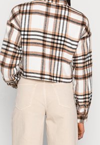 Krótka koszula flanelowa na guziki w biało-brązowo-czarną kratę. Posiada długie rękawy i kołnierz. Widać również dopasowane beżowe spodnie.