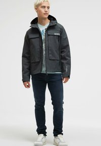 Wodoodporna czarna kurtka z kapturem, z dwoma przednimi kieszeniami i zapięciem na zamek, noszona na jasnoniebieskiej koszulce z grafiką i ciemnych jeansach.