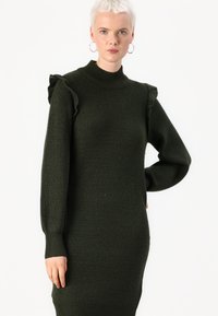 Robe pull côtelée vert foncé avec des manches longues et des accents de volants aux épaules, présentant un col haut et une silhouette ajustée.