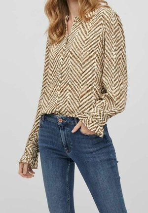 Femme portant un chemisier beige et marron à motif chevron avec des boutons, associé à un jean bleu taille haute, une main dans la poche.