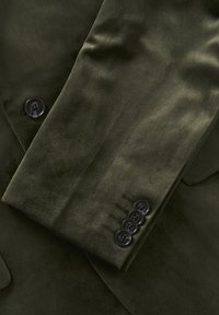 Sukně tmavě olivově zeleného blejzru s hladkou texturou, která má jeden černý knoflík a tři další knoflíky s dekorativním prošitím.