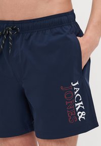 Marineblå svømmebukser laget av lett materiale med elastisk midje, snøring og en hvit og rød logo langs siden.