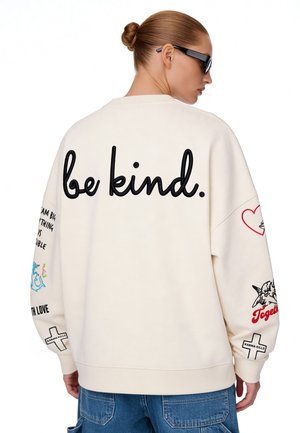 Person mit Dutt trägt ein weißes Sweatshirt mit „be kind.“ auf dem Rücken, Cartoon-Patches auf den Ärmeln und blaue Jeans.