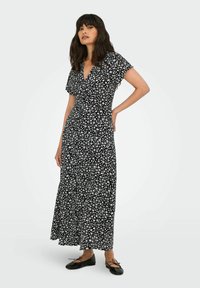 Zwarte maxi-jurk met bloemenmotief en korte mouwen, voorzien van witte bloemenpatronen, een V-hals en een vloeiende snit. Platte zwarte schoenen.