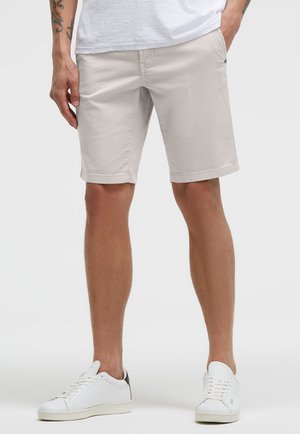 Mann trägt beige knielange Shorts, weiße Freizeitsneaker und ein weißes T-Shirt, mit teilweise in den Taschen steckenden Händen vor einem einfarbigen Hintergrund.