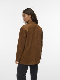 Mujer de cabello largo y oscuro que lleva un blazer de ante marrón y pantalones negros, mirando hacia la izquierda con un fondo blanco liso.