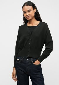 Zwarte cardigan met een V-hals, knoopsluiting en geribbelde zoom. Zachte textuur, losse pasvorm, gedragen over een zwarte top en donkerblauwe jeans.