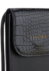 Czarny skórzany portfel z teksturą krokodylej skóry. Posiada złote logo "EXPATRIÉ", pleciony uchwyt zamka błyskawicznego oraz elegancki, kompaktowy design.