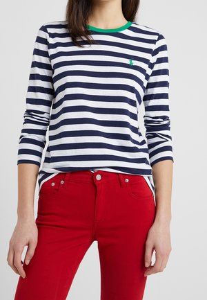 Kobieta ubrana w granatowo-białą, poziomo prążkowaną, długorękawową koszulę z zielonym kołnierzykiem oraz czerwone dopasowane spodnie, stoi na jednolitym tle.