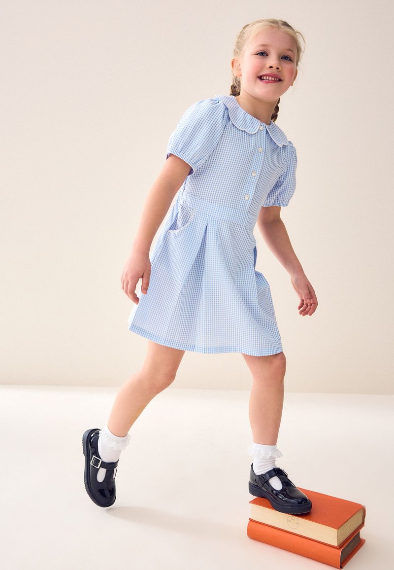 Jeune fille en robe bleue à carreaux, posant le pied sur deux livres orange empilés, avec des chaussures noires et des chaussettes blanches à froufrous, souriante et regardant devant elle.