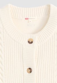 Cardigan tricoté couleur crème avec une texture côtelée, des détails en tricot torsadé et une fermeture à bouton au niveau du cou. L'étiquette indique "Levi's", taille S.