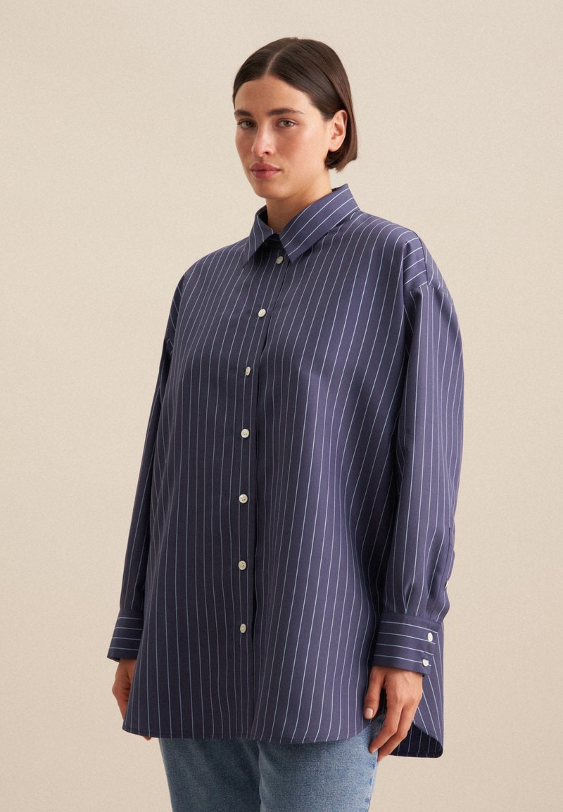 Camicia a maniche lunghe di colore blu navy con righe verticali bianche, colletto a bottoni, chiusura con bottoni e una vestibilità ampia e morbida. Tessuto di texture liscia.