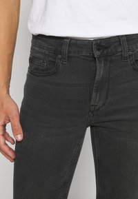 Krupan plan osobe u uskim crnim trapericama i bijeloj košulji, s lijevom rukom opuštenom uz tijelo.