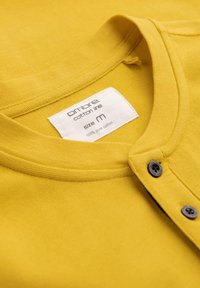Camicia di cotone gialla con colletto rotondo, due chiusure a bottone e un'etichetta che indica la taglia M e materiale 100% cotone puro.