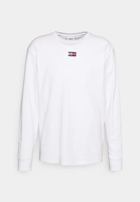 Weißes Langarmshirt mit Rundhalsausschnitt und kleinem Tommy Jeans Logo-Patch in Rot, Schwarz und Weiß, zentriert auf der Brust.
