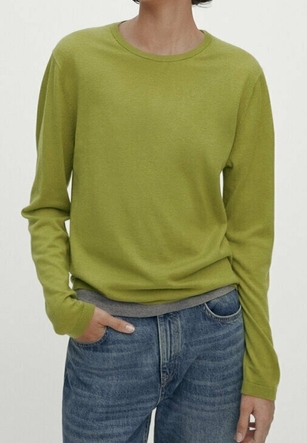 Zielony sweter z długim rękawem i okrągłym dekoltem, wykonany z miękkiego materiału. Połączony z niebieskimi dżinsami, z widoczną szarą warstwą pod spodem na wysokości talii.