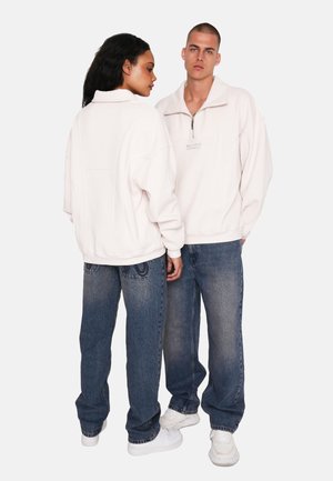Lyskinnende pink pullover med høj krave og kvart lynlås, parret med løse blå denimjeans. Har subtil branding på forsiden.