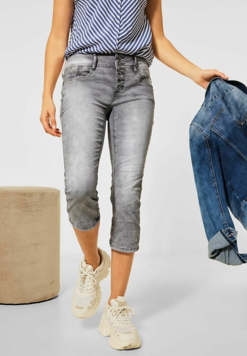 Woman wearing gray cropped jeans and white sneakers, holding a blue denim jacket in her right hand.