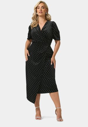 Femme portant une robe noire mi-longue avec un motif losange argenté, des manches bouffantes et des sandales argentées à talons hauts, debout devant un fond uni.