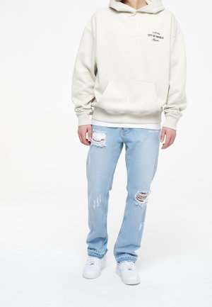 Ljusbeige hoodie med en framficka och svart text. Ljusblå slitna jeans med revor. Vita sneakers fulländar outfiten.