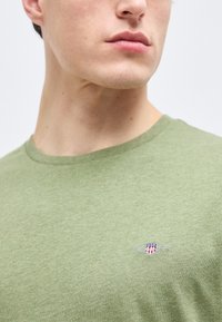 Νεαρός άνδρας με ανοιχτό πράσινο t-shirt με στρογγυλή λαιμόκοψη και μικρό κέντημα με κόκκινη, λευκή και μπλε ασπίδα στο στήθος.