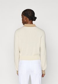 Μπεζ πλεκτό πουλόβερ με μακριά μανίκια και ριμπ τελείωμα. Διαθέτει καφέ γιακά με λευκές ρίγες, που αναδεικνύει ένα απαλό, υφασμάτινο ύφασμα με υφή.