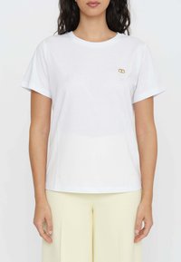 T-shirt bianco in cotone con maniche corte e scollo tondo, caratterizzato da un piccolo logo dorato sul petto. Tessuto liscio, vestibilità rilassata.