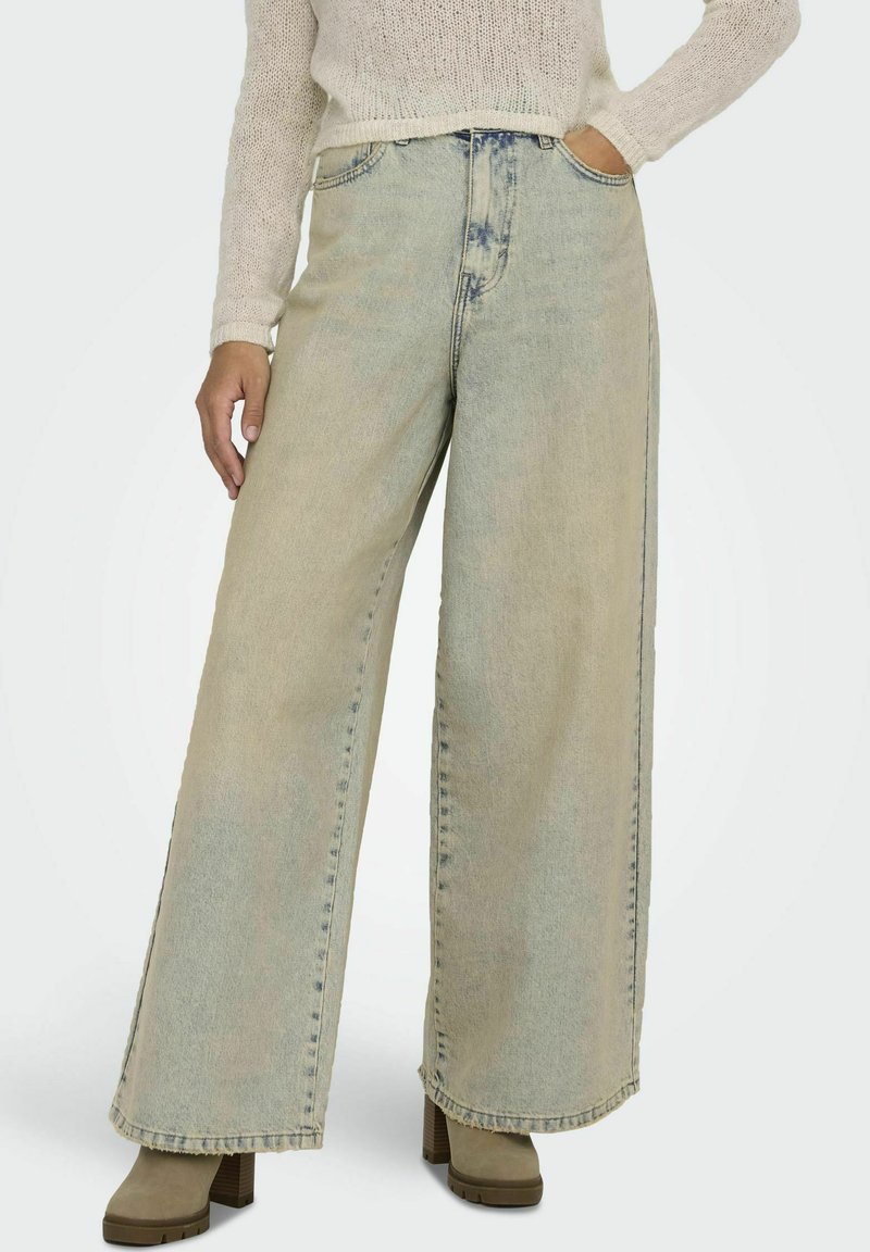 Jasnoniebieskie spodnie dżinsowe w stylu wide-leg, wykonane z denimu z wyblakłym wykończeniem, z surowym brzegiem i widocznym szwem. Połączone z masywnymi, beżowymi botkami na wysokim obcasie.
