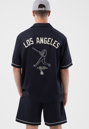 Uomo di spalle che indossa una maglietta blu navy e pantaloncini con bordi bianchi; sulla maglietta è presente la scritta "LOS ANGELES", una grafica di un giocatore di baseball e il logo "LA".