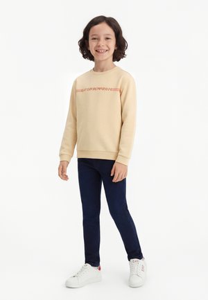 Niño con cabello rizado, de pie y sonriendo, vestido con una sudadera beige con letras rojas, pantalones oscuros y zapatillas blancas.