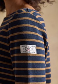 Personne portant un t-shirt à manches longues rayé horizontalement bleu marine et beige avec une étiquette blanche rectangulaire sur le haut du bras.
