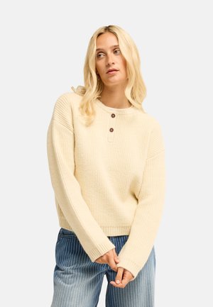 Donna bionda che indossa un maglione a coste color crema con bottoni e pantaloni blu a righe sottili, guarda leggermente di lato con le mani giunte.