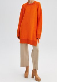 Przewymiarowany, pomarańczowy sweter w prążki z długimi rękawami i okrągłym dekoltem, zestawiony z beżowymi spodniami o szerokich nogawkach oraz beżowymi botkami na szerokim obcasie.