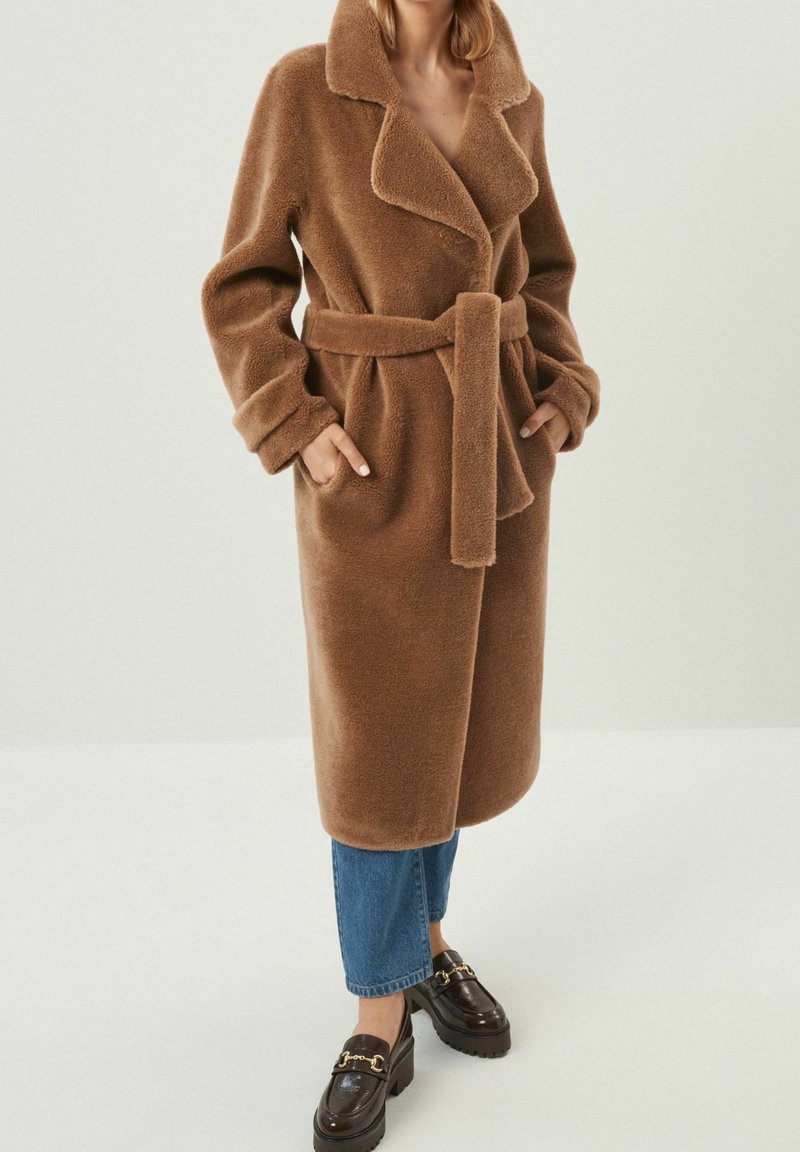 Femme portant un long manteau marron pelucheux ceinturé, un jean bleu court et des mocassins marron foncé avec des détails en chaîne dorée.