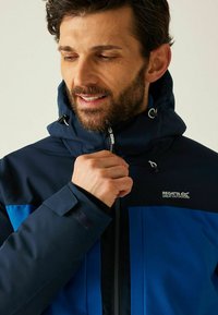 Homme barbu fermant une veste d'extérieur bleu marine et bleu avec le logo "Regatta Great Outdoors" sur la poitrine, mettant en avant les détails de la veste.