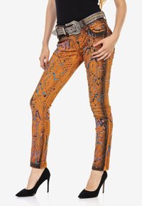 Jeans oranges avec un design texturé, ornés de motifs éclaboussés bleus et violets. Détails avec une ceinture argentée ornée de bijoux et des talons hauts.