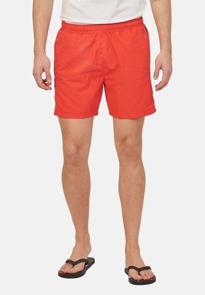 Hombre con pantalones cortos de baño rojo brillante, una camiseta blanca y chanclas negras, de pie frente a un fondo liso.