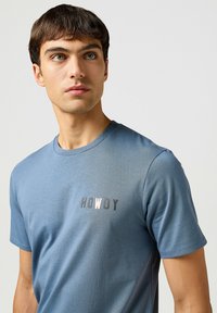 Camiseta de manga corta azul hecha de tela suave. Presenta un destacado estampado de "HOWDY" en tonos oscuros y claros en el lado izquierdo del pecho.