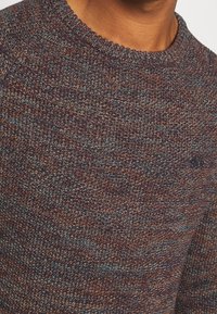 Strickpullover mit einem melierten Muster in Dunkelbraun, Blau und Rostfarben. Gerippter Rundhalsausschnitt mit einem dezenten Logo-Detail.