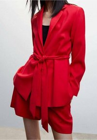 Conjunto de blazer rojo y pantalones cortos, confeccionado en tela suave. El blazer cuenta con cinturón en la cintura, solapas con muesca y bolsillos laterales.