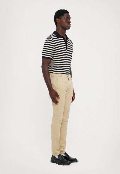 Uomo in piedi di lato, vestito con una polo a maniche corte a righe bianche e nere, pantaloni beige, calze bianche e mocassini neri, su uno sfondo neutro.
