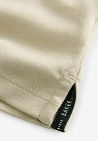 Beige bomullsskjorta i hörnet med synlig sömnad och en svart etikett märkt "BAKER", som visar en slät textur och rena kanter.